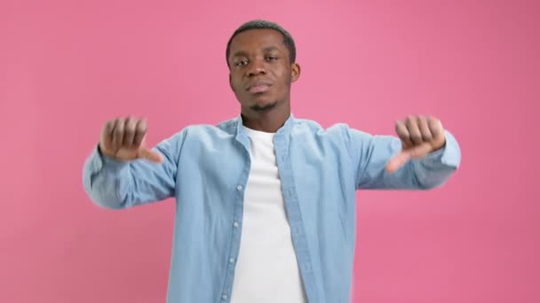 Ontevreden jonge zwarte man Afro-Amerikaanse in denim shirt en wit T-shirt toont duimen naar beneden, afkeer, negatieve feedback afkeer, slechte feedback, emotie van niet-goedkeuring geïsoleerd op roze achtergrond. — Stockvideo
