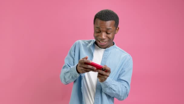 Gokken jonge Afro-Amerikaanse man in blauw denim shirt maakt gebruik van racen op mobiele telefoon, houdt smartphone gadget voor PC, video games geïsoleerd op roze studio achtergrond. Emotie winnen op video game. — Stockvideo