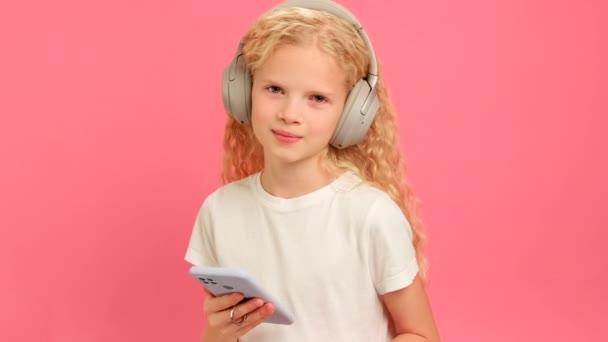 Ξανθιά Καυκάσια έφηβη απολαμβάνει να ακούει μουσική με ακουστικά Bluetooth στο κεφάλι της, να χορεύει και να ακούει το αγαπημένο της τραγούδι στο τηλέφωνό της σε ροζ φόντο στούντιο. — Αρχείο Βίντεο