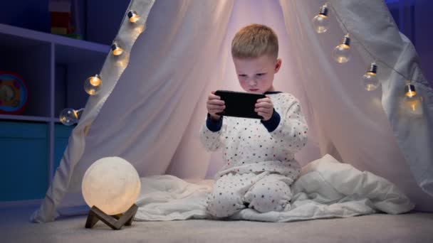 하얀 잠옷을 입은 작은 금발의 소년 이 밤중에 스마트폰에서 경기를 하고 있는 현란 한 구근으로 장식된 손으로 만든 텐트 위에 앉아 있다. 밤에 스마트폰으로 레이싱하는 아이들. — 비디오