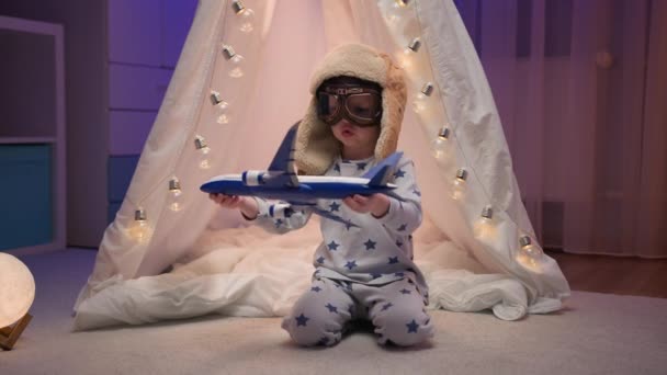 Niño en pijama, gafas y un sombrero de aviapilot está sentado en habitación oscura con carpa blanca hecha a mano, wigwam con luces brillantes y jugando con el avión de juguete. Niño vuela en avión, deseo de volar. — Vídeos de Stock