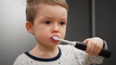 Küçük çocuk dişlerini diş fırçası ve diş fırçasıyla aynanın önünde duran bakkaldan ve plaketten temizliyor. Oral hijyenle meşgul, ayakta duran ve aynaya bakan sarışın çocuk portresi..