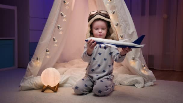 Маленький хлопчик грає з іграшковим білим літаком, мріє літати і бути капітаном літака, який сидить у темній кімнаті біля намету з сяючими гірляндами. Мрії про дитину літати, концепція подорожі авіаперельоти . — стокове відео