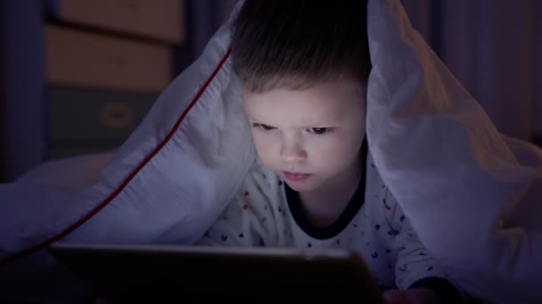 Retrato rubio pequeño niño de 4 años cubierto con manta blanca se encuentra en el suelo y mira la pantalla brillante tableta, ve videos y juega en el ordenador portátil por la noche en el dormitorio. Navegar por Internet. — Vídeo de stock