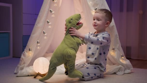 Mały blondyn bawi się swoją pluszową zabawką dinozaura, przytulając ją i przytulając do klatki piersiowej, radując się szczęściem siedząc w domu w nocy w pobliżu ręcznie robionego namiotu. Dziecko kocha zabawki. — Wideo stockowe