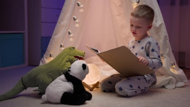 Menino loiro de pijama lendo conto de fadas para seu brinquedo de panda recheado e dinossauro verde sentado confortavelmente ao lado da tenda no quarto escuro da noite. Conceito de amor, infância, amizade, imaginação, criatividade — Vídeo de Stock