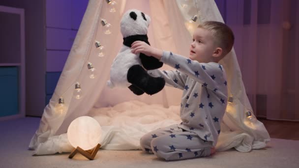 Kleine blonde jongen speelt met zijn gevulde panda speelgoed, knuffelt haar en knuffelt haar op zijn borst, verheugen zich met geluk terwijl thuis zitten 's nachts in de buurt van handgemaakte tent. Kinderliefde voor zacht speelgoed. — Stockvideo