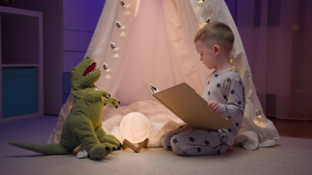 Fotografía cinematográfica auténtica del niño rubio leyendo cuento de hadas su juguete tiranosaurio rex felpa, cómodamente sentado en la tienda de campaña en el dormitorio de la noche. Concepto amor, infancia, amistad, imaginación y creatividad. — Vídeos de Stock