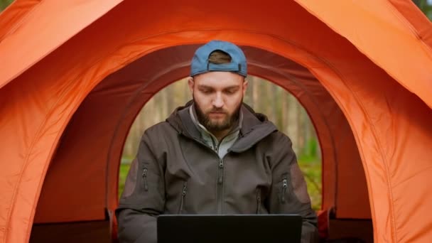 Moving camera back, portrait male freelance tourist remotely working at computer, sitting in an orange tent in nature in woods, drinking hot coffee and working on laptop. Outdoor work with laptop. — Stock Video