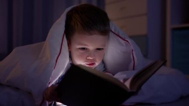 5 yaşındaki küçük sarışın beyaz çocuk, beyaz battaniyeyle kaplı, karanlık yatak odasında uzanıyor, peri masalları ve gece lambasından gelen ışıkla kitap okuyor. Küçük çocuk akşamları evde kitap okuyor..
