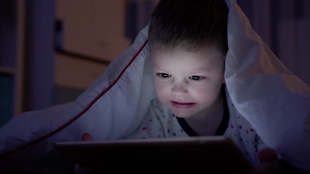 Retrato pequeño niño rubio experimenta la emoción del miedo y la ira mientras ve películas de miedo o dibujos animados en la tableta, acostado noche bajo una manta blanca en la noche en el suelo en casa. experimenta emociones miedo. — Vídeo de stock