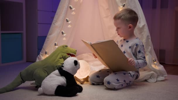 かわいいブロンドの男の子は テントの近くの夜に家に座っている間 友人のテディベアパンダと緑の恐竜のレックスに就寝時の物語を読み取ります 幼馴染 — ストック動画