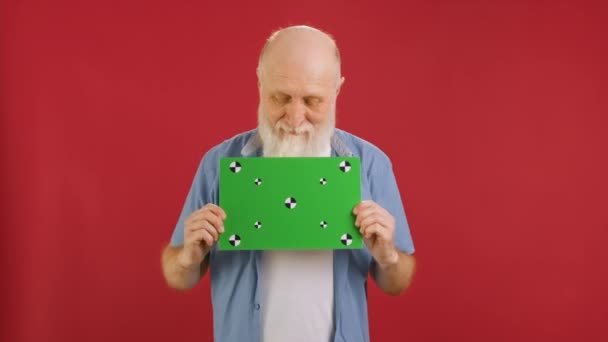 Ευτυχισμένος γέρος κρατώντας Banner με πράσινο Screen Tracks σημεία για Copy Space. Κενό πράσινο πίνακα οθόνης. Έκπληκτος καλύπτει το στόμα του με το χέρι του με χώρο για κείμενο ή διαφήμιση σε κόκκινο φόντο. — Αρχείο Βίντεο