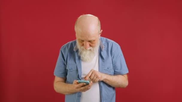 Choquée heureuse victoire, vieux barbu grand-père 60 ans en chemise bleue, émotion de gagner au casino ou paris sportifs dans l'application mobile pose en isolement sur fond de studio rouge. — Video