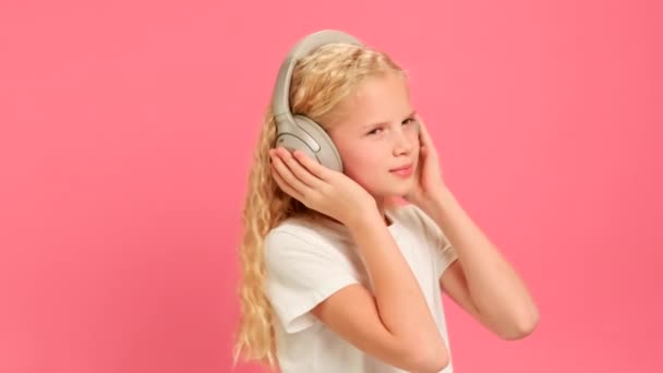 慢动作肖像女孩喜欢用耳机听歌曲。穿着灰色耳机听着自己最喜欢的音乐，并在粉红工作室背景下看着摄像机的金发少女形象. — 图库视频影像