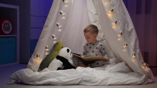 Menino loiro de pijama lê conto de fadas para seus brinquedos de panda recheados e rex dinossauro verde sentado confortavelmente na tenda branca no quarto escuro da noite. Conceito de amor, infância, amizade, criatividade. — Vídeo de Stock