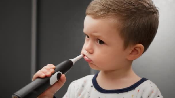Portrait blond boy engaged in oral hygiene, standing and looking in mirror. Little boy cleans his teeth from caries and plaque with an electric toothbrush with toothpaste, standing in front of mirror. — Stock Video