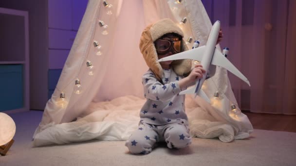 子供は飛行機のおもちゃの欲望のフライで飛ぶ。白い手作りのテントで暗い部屋に座っているパジャマ、眼鏡、飛行士の帽子の小さな男の子、光るライトでウィグワム、おもちゃの飛行機で遊ぶ. — ストック動画
