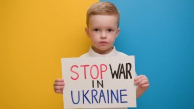 Küçük sarışın çocuk Ukrayna 'daki savaşı durdurmak için çağrıda bulunuyor, Ukrayna' da mavi-sarı stüdyo arka planında duran savaşı durdurmak için yazıyla bayrağı kaldırıyor. Savaş yok, savaşı durdurun, Rus saldırıları.