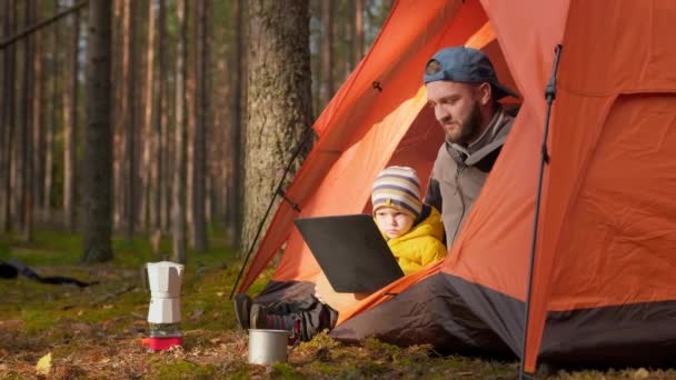 Батько і син відпочивають разом у природі у весняному лісі, сидячи в помаранчевому наметі і переглядаючи відео разом на екрані ноутбука, спілкуючись через відео чат через ноутбук з родичами . — стокове відео