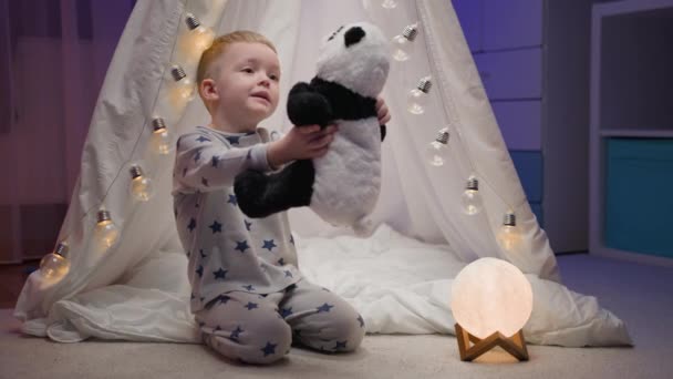 Menino loiro está brincando com seu brinquedo de panda recheado, abraçando-a e abraçando-a ao peito, regozijando-se com a felicidade enquanto se senta em casa à noite perto de tenda artesanal. Amor infantil para brinquedos macios. — Vídeo de Stock