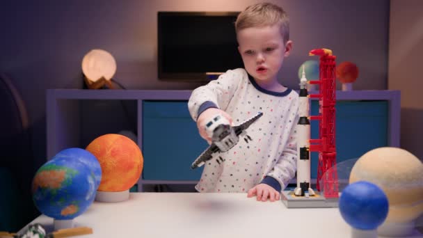 Petit garçon blond enfant joue avec la navette spatiale du constructeur, vole près des planètes du système solaire en soirée. Enfant s'imagine pilote de vaisseau spatial. Voler dans l'espace, voler sur un vaisseau spatial. — Video