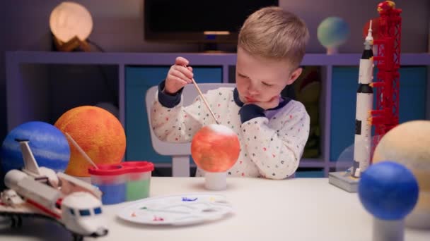 Blondine jongen schilderen planeet Mars van het zonnestelsel zitten aan tafel in de avond met planeten van het zonnestelsel en ruimte raket van constructeur geplaatst op tafel. Cosmonauticadag op 12 april. — Stockvideo