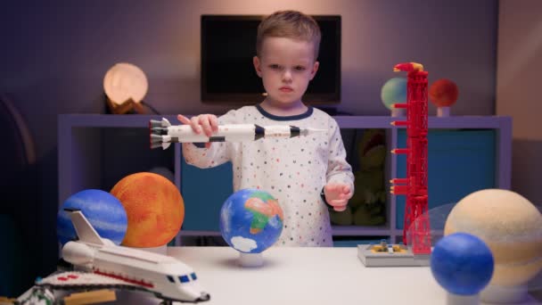 Kleine blonde jongen speelt met ruimteschip van constructeur, vliegt in de buurt van planeten van het zonnestelsel in de avond. Kind stelt zich voor als piloot van een ruimteschip. Verlangen vliegen in de ruimte, vliegen op ruimteschip. — Stockvideo