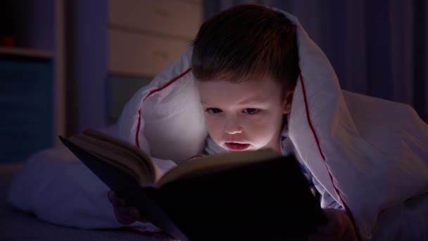 Маленький белокурый кавказский мальчик 5 лет лежит на полу, покрытом белым одеялом в темной спальне, читая книгу со сказками и светом от ночной лампы. Маленький мальчик изучает книгу вечером дома. — стоковое видео
