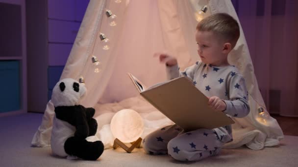 Χαριτωμένο ξανθό 4 ετών μικρό παιδί διαβάζει βιβλίο για το αγαπημένο του παιχνίδι panda αρκούδα, gesticulating και κουνώντας το δάχτυλό του, ενώ κάθεται σε σκοτεινό δωμάτιο με λευκή σκηνή διακοσμημένα με λαμπερό γιρλάντες. — Αρχείο Βίντεο