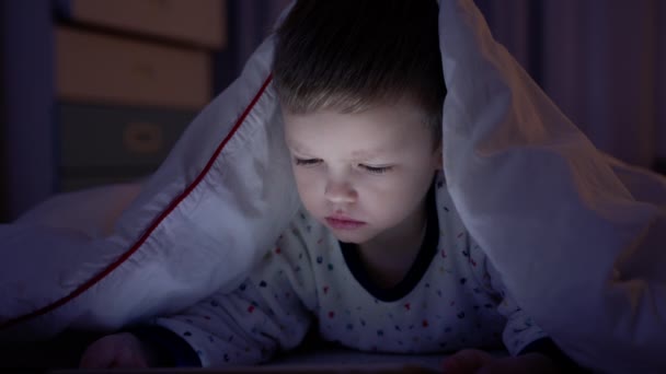 Retrato pequeño niño rubio experimenta la emoción del miedo y la ira mientras ve películas de miedo o dibujos animados en la tableta, acostado noche bajo una manta blanca en la noche en el suelo en casa. experimenta emociones miedo. — Vídeo de stock