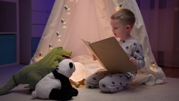 Menino loiro de pijama lendo conto de fadas para seu brinquedo de panda recheado e dinossauro verde sentado confortavelmente ao lado da tenda no quarto escuro da noite. Conceito de amor, infância, amizade, imaginação, criatividade — Vídeo de Stock