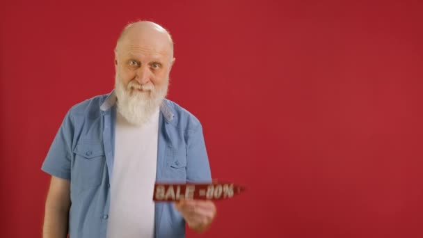 Old bearded man Holds Sign With Inscription Sale -80 off, Points His Finger at Empty Space Mock up and Looks at Discount in Camera and Smiles on Red Background. Μεγάλες εκπτώσεις τις μαύρες Παρασκευές. — Αρχείο Βίντεο