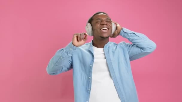 Ευτυχισμένος μαύρος έφηβος 20 χρόνια, σε denim πουκάμισο με ακουστικά στο κεφάλι του ακούει την αγαπημένη του μουσική, τραγουδά λόγια τραγουδιού, κουνάει τα χέρια του στο ρυθμό της μουσικής σε ροζ φόντο στούντιο. — Αρχείο Βίντεο