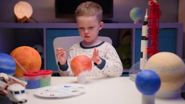 Blond boy schildert planeet zonnestelsel Mars met kleurrijke verven zitten thuis tafel in de avond, planeten zonnestelsel, ruimteschepen en spaceshuttle van constructeur rond. Cosmonauticadag op 12 april. — Stockvideo