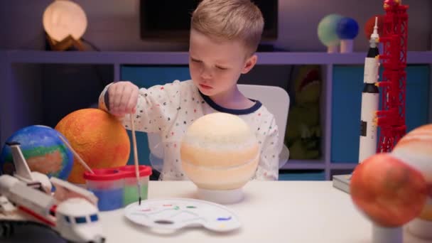 Biondo ragazzo dipingere pianeta sistema solare Saturno con vernice colorata seduto a casa tavolo in serata, pianeta sistema solare, astronavi e navetta spaziale dal costruttore intorno. Giorno di Cosmonautica il 12 aprile. — Video Stock
