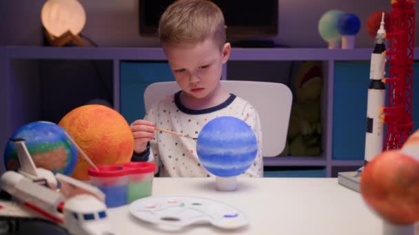 Loiro menino pintar planeta sistema solar Neptune com pintura colorida sentado em casa mesa à noite, planeta sistema solar, naves espaciais e ônibus espacial do construtor ao redor. Dia da Cosmonáutica em 12 de abril. — Vídeo de Stock