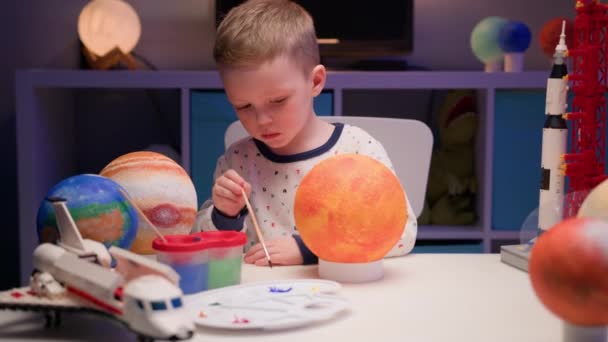 ブロンドの少年は、夜に家のテーブルに座って、太陽系の惑星、宇宙船や周りのコンストラクタからのスペースシャトルで太陽の星を描きます。宇宙の日は4月12日. — ストック動画