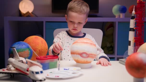 Blond boy verf planeet zonnestelsel Jupiter met kleurrijke verf zit thuis tafel in de avond, planeet zonnestelsel, ruimteschepen en spaceshuttle van constructeur rond. Cosmonauticadag op 12 april. — Stockvideo