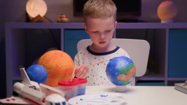 Blond boy verf planeet zonnestelsel Aarde met kleurrijke verf zittende thuis tafel in de avond, planeet zonnestelsel, ruimteschepen en spaceshuttle van constructeur rond. Cosmonauticadag op 12 april. — Stockvideo