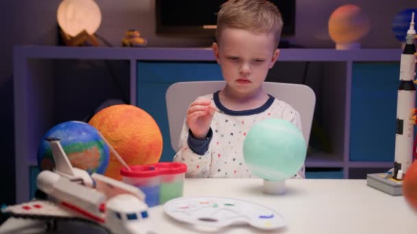 Loira menino pintar planeta sistema solar Urano com tinta colorida sentado em casa mesa à noite, planeta sistema solar, naves espaciais e ônibus espacial do construtor ao redor. Dia da Cosmonáutica em 12 de abril. — Vídeo de Stock
