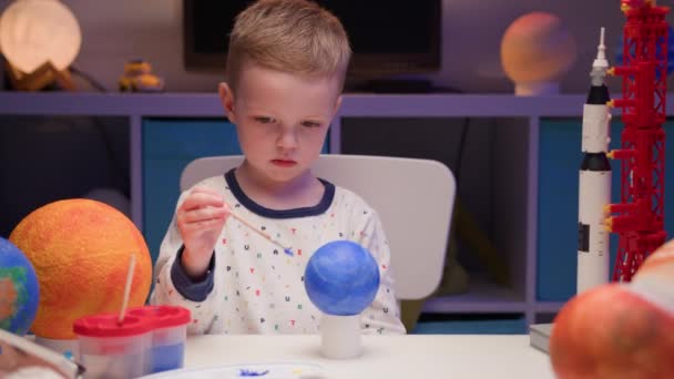 Blond boy verf planeet zonnestelsel Pluto met kleurrijke verf zit thuis tafel in de avond, planeet zonnestelsel, ruimteschepen en spaceshuttle van constructeur rond. Cosmonauticadag op 12 april. — Stockvideo