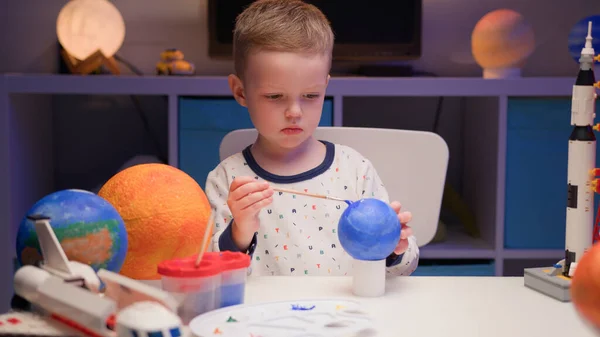 ブロンドの少年は、夜に家のテーブルに座ってカラフルな塗料で惑星の太陽系冥王星をペイント,惑星の太陽系,周りのコンストラクタから宇宙船やスペースシャトル.4月12日の宇宙の日. — ストック写真