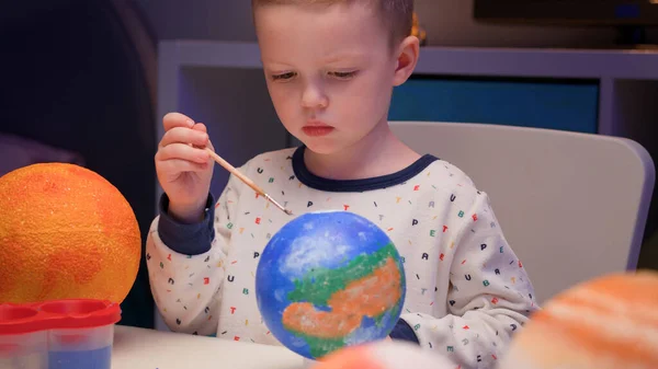 Blond boy paint planet solar system Earth with colorful paint sitting home table in evening, planet solar system, spaceships and space shuttle from constructor around. Cosmonautics Day on April 12. — Stock Photo, Image