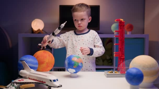 Kleine blonde jongen speelt met ruimteschip van constructeur, vliegt in de buurt van planeten van het zonnestelsel in de avond. Kind stelt zich voor als piloot van een ruimteschip. Verlangen vliegen in de ruimte, vliegen op ruimteschip. — Stockvideo
