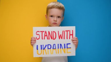 Portre Sarışın çocuk Ukrayna 'nın yanında durmak için sesleniyor, yazıyla bayrağı kaldırıyor Ukrayna' daki savaşı durdurun mavi-sarı stüdyo arka planında duruyor. Savaş yok, savaşı durdurun, Rus saldırıları.