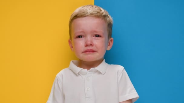 Portret kleine blonde jongen huilen op geel-blauwe achtergrond vlag van Oekraïne. Crisis in Oekraïne, oorlog tegen kinderen, Russische agressie humanitaire ramp, Hulp aan kinderen in Oekraïne. — Stockvideo