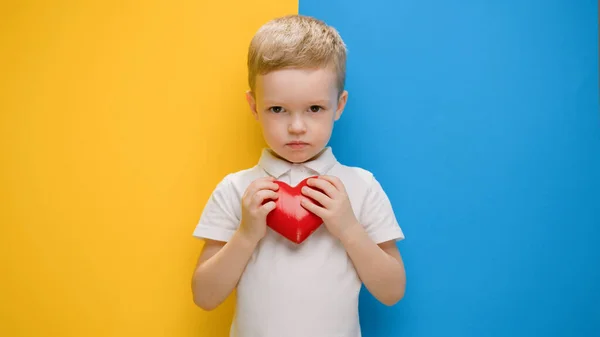 Little blond boy expresses love, presses red heart with his hands to his chest, standing on blue-yellow background. Call to love, hands in shape of heart, Ukraine with love. Child is pacifist. — Stock Photo, Image