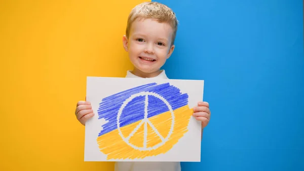 Blond-haired boy smiling on yellow-blue background of flag of Ukraine holds banner with logo of anti-war movement Pacific. Pacifist child protests against war in Ukraine. — Stock Photo, Image