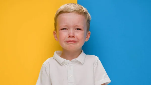 Portrét malý blonďatý chlapec pláče na žlutomodré pozadí vlajky Ukrajiny. Krize na Ukrajině, válka proti dětem, ruská agrese, humanitární katastrofa, Pomoc dětem na Ukrajině. — Stock fotografie